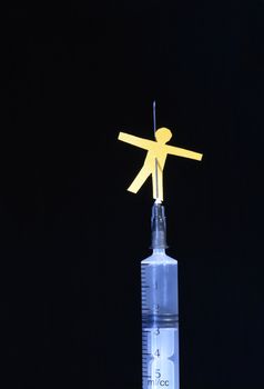 Yellow paper man pierced with needle of syringe on dark background