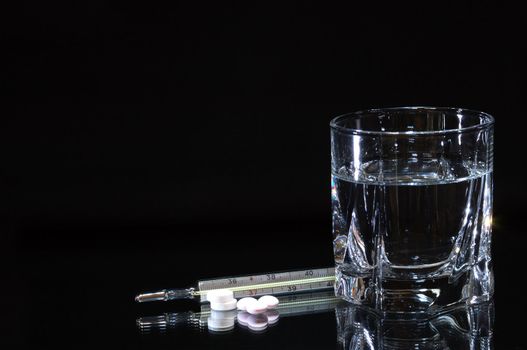 Few pills and thermometer lying near glass of water isolated on dark background with copy space