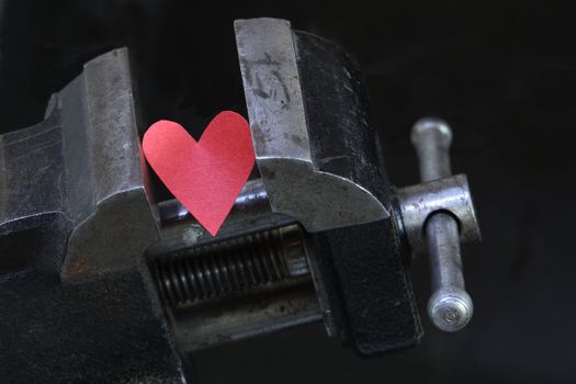 Small red paper heart under pressure with old vise grip on dark background