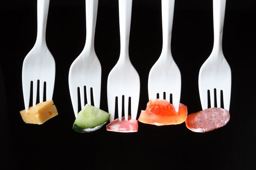 Five white plastic forks in a row with various food on a black background