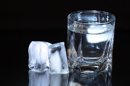 Two ice cubes near glass of water isolated on dark background