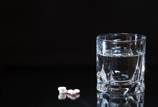 Few pills lying near glass of water isolated on dark background with copy space