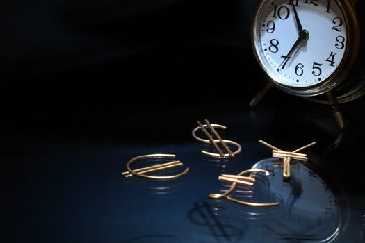 World currency signs made from brass wire near clock on dark background with copy space