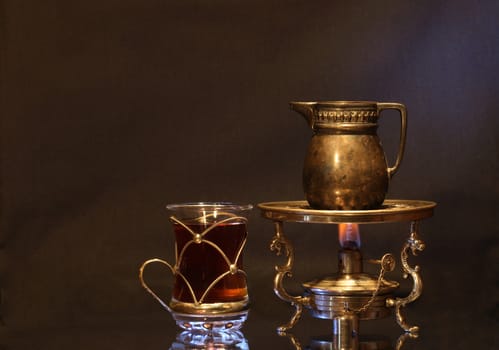 Cup of tea near ancient brass jug standing on vintage table spirit lamp with flame