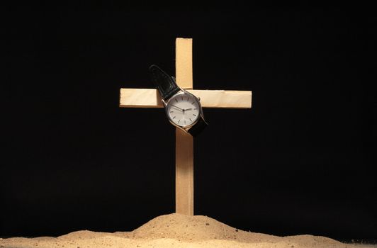 Watch hanging on wooden cross standing in sand on black background