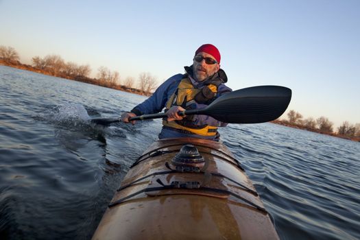 mature male paddler exercising (turning boat using rudder stroke with his wing carbon fiber paddle) in a home built wooden sea kayak on lake, fall scenery  in Colorado, view from kayak bow