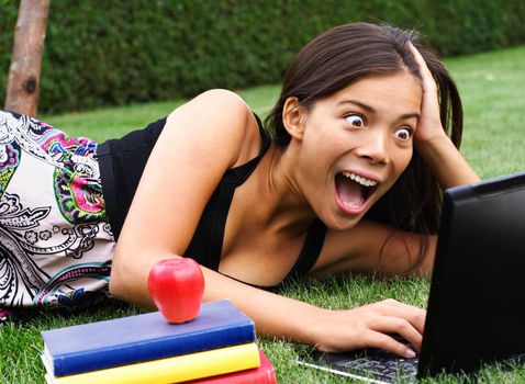 Student with laptop surprised while chatting / checking email / reading gossip news in the park. Beautiful mixed race caucasian / asian model.