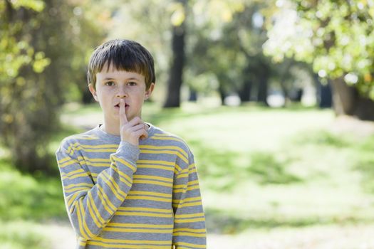 Cute Young Boy Standing Outdoors in a Park  With Finger Up To Mouth