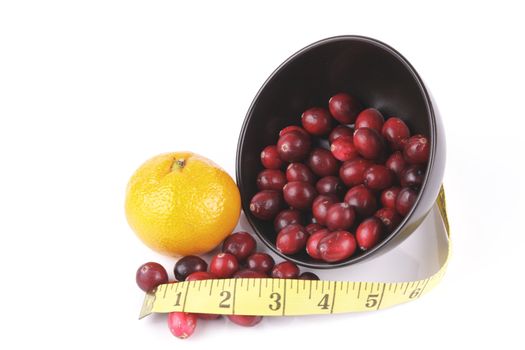 Red ripe cranberries spilling out of a small round black bowl on its side with a tape measure and satsuma on a reflective white background