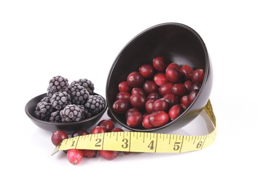 Red ripe cranberries spilling out of a small round black bowl on its side with a tape measure and frozen blackberries on a reflective white background