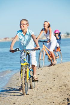 Cute girl with her mother and brother ride bikes along the beach. Focus on girl. Vertical view