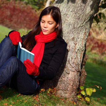 Woman reading book and relaxing outdoors in park under tree in autumn.