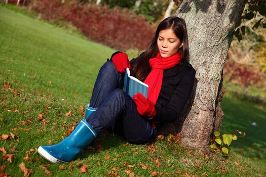  Woman reading book outdoors in park under tree in autumn.