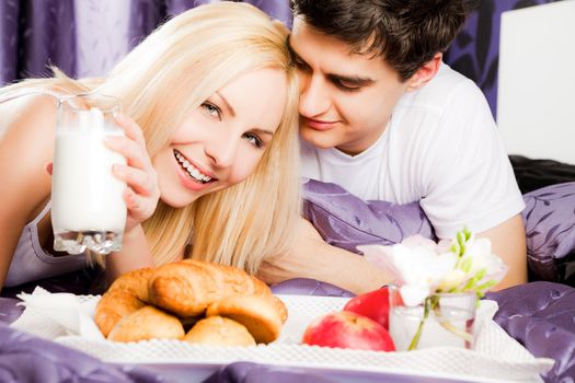 Happy young couple lying and having breakfast in bed, woman looking at camera