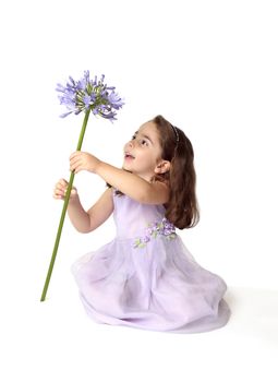 A little girl sitting on the floor in pretty mauve dress decorated with floral appliques, is spinning a large stemmed agapanthus lily flower in bloom, with much delight and excitement.   