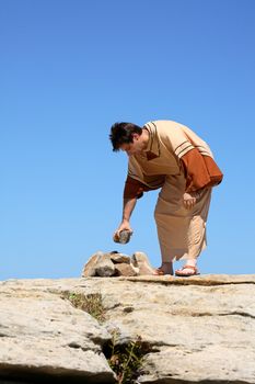 Full length man in brown and beige  tunic robe bending over near a pile of rocks.  Concept sin, law or culture.  This form of punishment was used in many ancient cultures and is still used in some cultures today.   Space for a heading, title or verse.