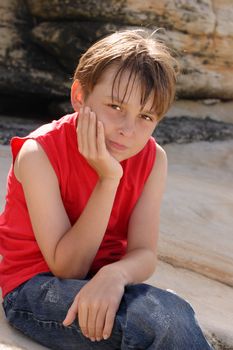 A young child dressed in jeans and tank top sitting and thinking by rocks.