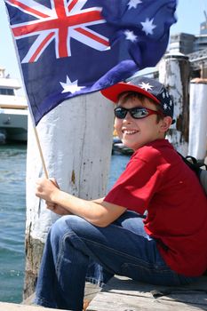 Child relaxing on harbourside wooden pier smiles happily and is waving an Australian flag.