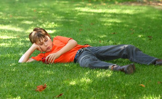 a young boy sprawled out on the shaded grass in the park, listens to music on an mp3 music player.