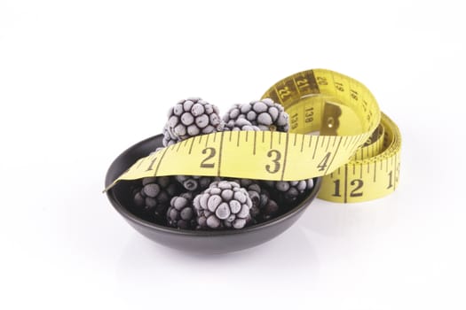 Black ripe frozen blackberries in a small round black dish with a tape measure on a reflective white background