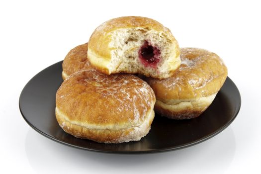 Black round plate of jam doughnuts with a bite mark on one showing red jam on a reflective white background