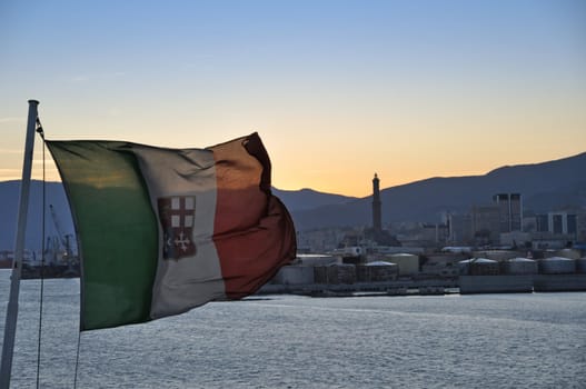 An Italian flag on a ferry leaving the port of Genova. The lighthouse Lanterna on the sunset background