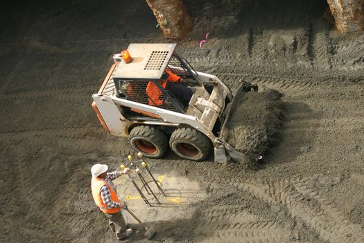 A bobcat shovels gravel while another workman ensures the site markings are kept clear.
