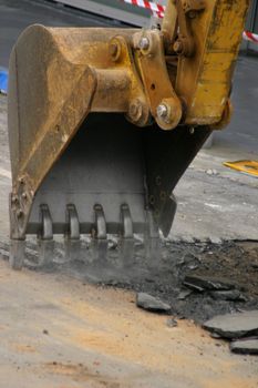 Large steel teeth of the excavator vibrate and shake noisily as they begin to dig up an existing road.
