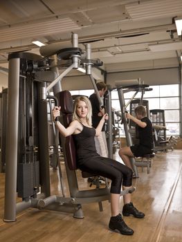 Girl using an exercise machine at a health club with two men in the background