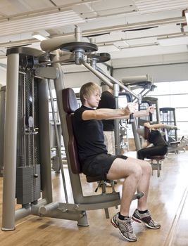 Young man using an exercise machine at a health club with a girl and a boy in the background