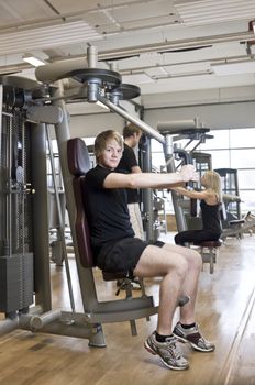 Young man using an exercise machine at a health club with a girl and a boy in the background