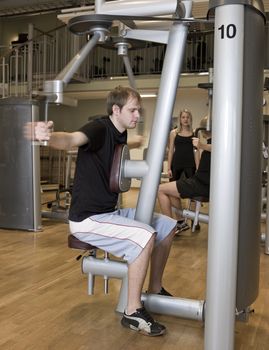 Young man using an exercise machine at a health club with a girl and a boy in the background