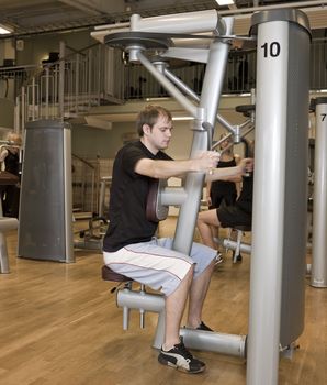 Young man using an exercise machine at a health club with a girl and a boy in the background