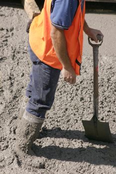 This construction worker stands in wet cement with knee high boots  covered in cement.  He is leaning on his his shovel
