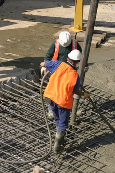 Workers standing on thick steel mesh and pouring concrete through a large hose attached to a long extendable boom connected to a concrete hydraulic truck mounted pump..   There is motion in the pouring cement.
