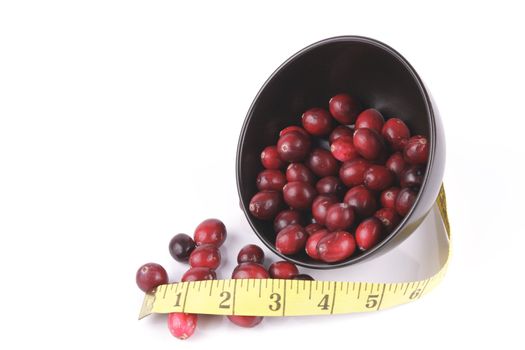 Red ripe cranberries spilling out of a small round black bowl on its side with a tape measure on a reflective white background