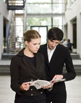 Two women reading a magazine while standing in an office building