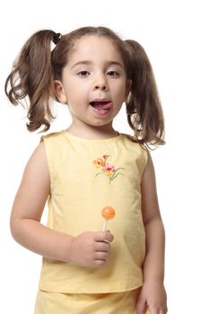 A little girl licks her lips.  She iis wearing a yellow outfit with hair in ponytails and she is holding a candy lollipop