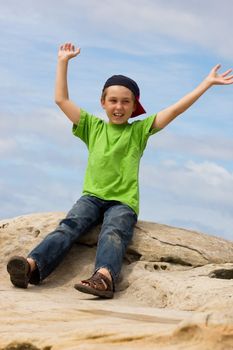 Carefree child playing waving arms gregariously overhead.  He is sitting on a rock against a pretty blue sky