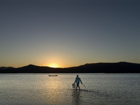 An unrecognizable fisherman walks through the shallow lake to reach his boat on the magic hour.