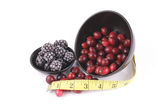 Red ripe cranberries spilling out of a small round black bowl on its side with a tape measure and frozen blackberries on a reflective white background