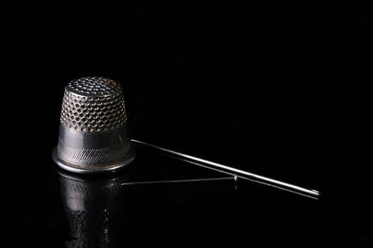 Thimble for protection of a finger during sewing, needles and a pin on a black brilliant surface.