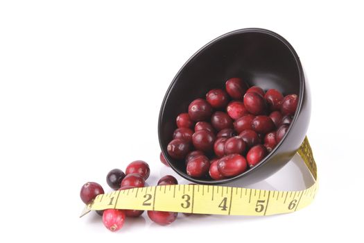Red ripe cranberries spilling out of a small round black bowl on its side with a tape measure on a reflective white background