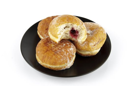 Black round plate of jam doughnuts with a bite mark on one showing red jam on a reflective white background