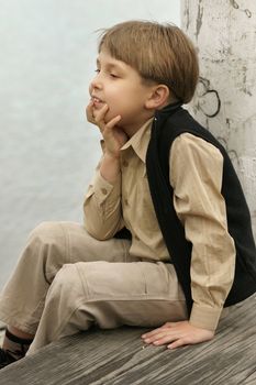 Young boy sitting on weathered timber leaning on a pylon near the water.