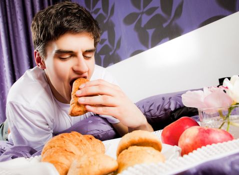 Young male having breakfast in bed, biting croissant