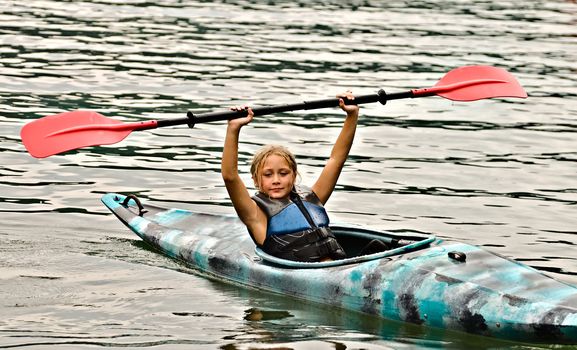 A cute young girl in a kayak holding a paddle above her head giving a signal.