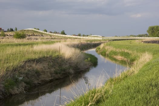 windy irrigation ditch with flowing water, green meadows and pasture behind  white fence, northern Colorado
