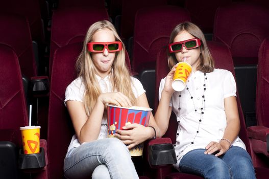 two girls look three-dimensional cinema, sitting in the glasses, eat popcorn, drink drink