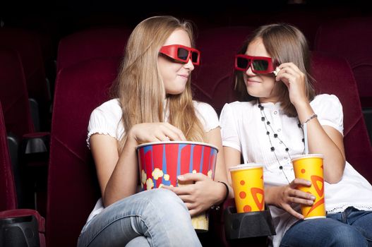 two girls look three-dimensional cinema, sitting in the glasses, eat popcorn, drink drink